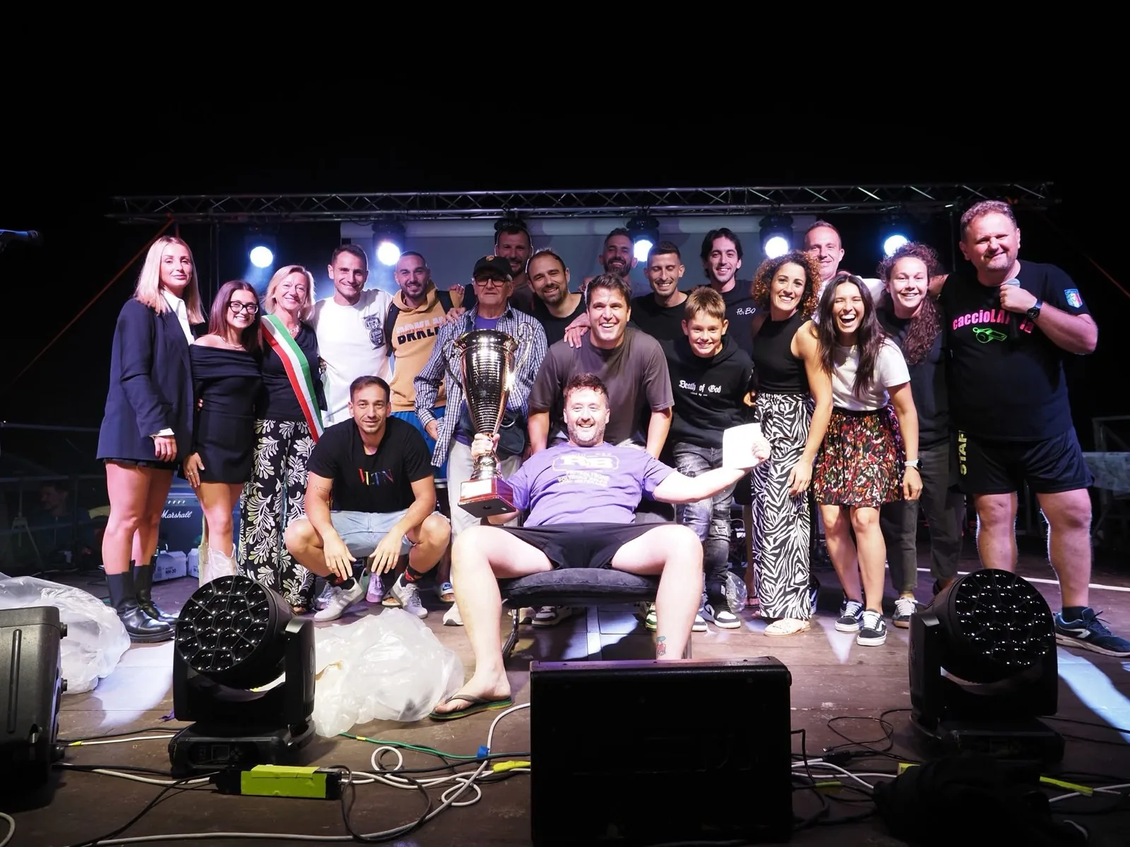 La premiazione del Calcetto Saponato 2025: il vincitore seduto con la coppa, circondato dai compagni, dallo staff del comitato e dalle autorità locali sul palco della festa.